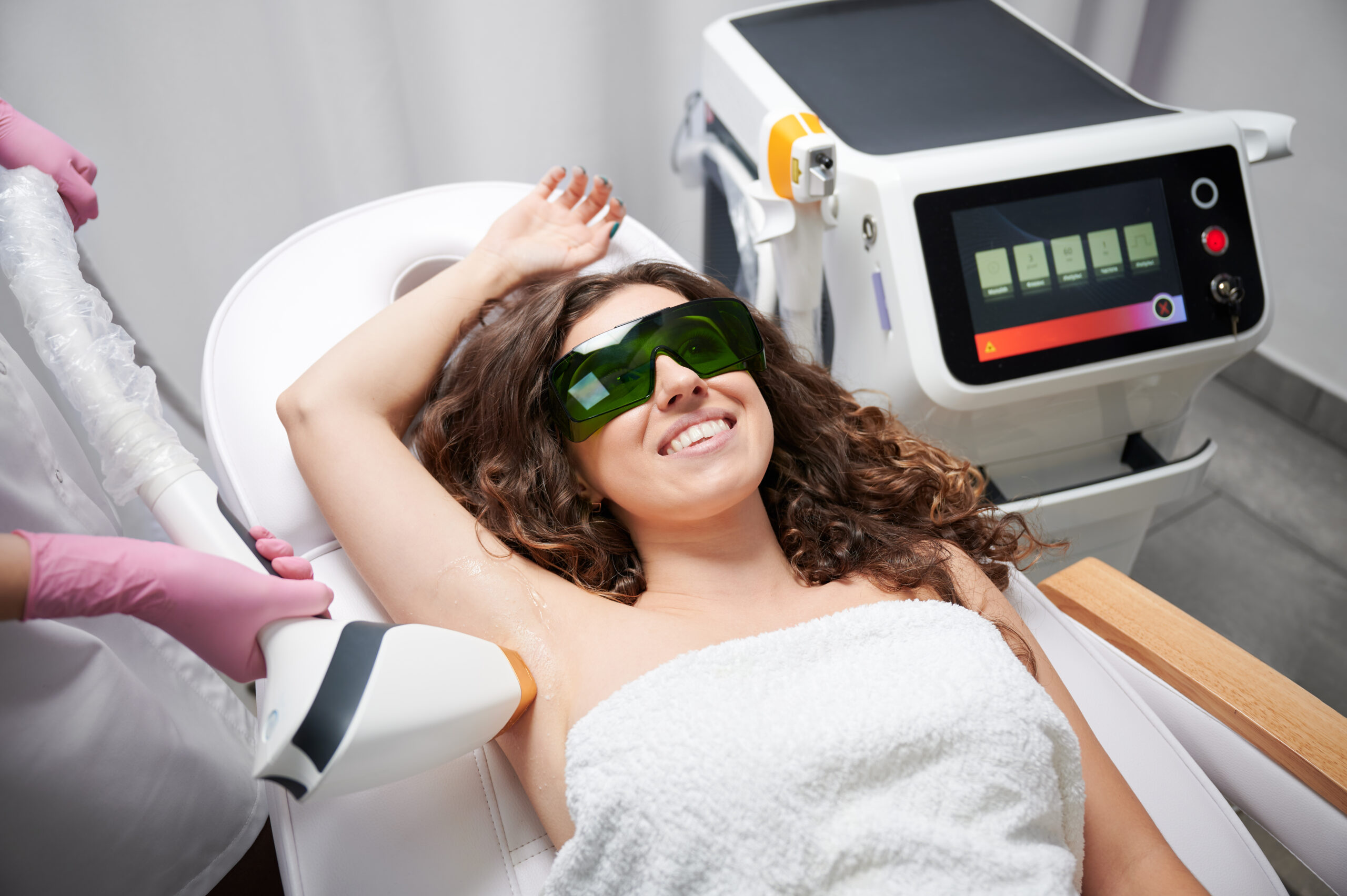 Young woman in safety glasses lying on daybed and smiling while beautician removing hair from female armpit with laser device. Cosmetologist performing laser hair removal procedure in beauty salon.