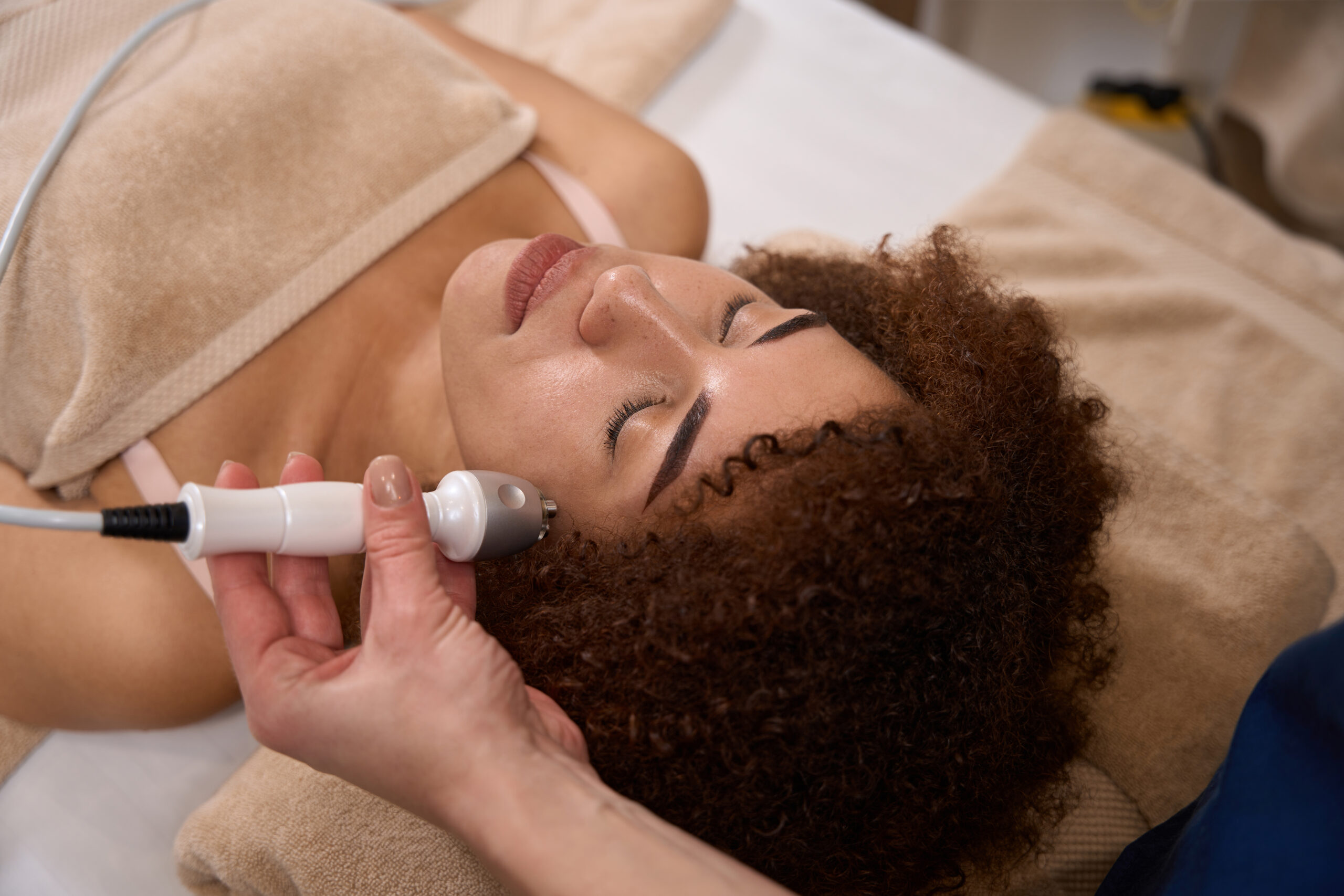 African American woman is lying on a couch in a spa salon and a specialist is doing a RF lifting procedure for her face